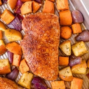 A seasoned baked chop on a tray surrounded by roasted sweet potatoes, apples, and red onion pieces.