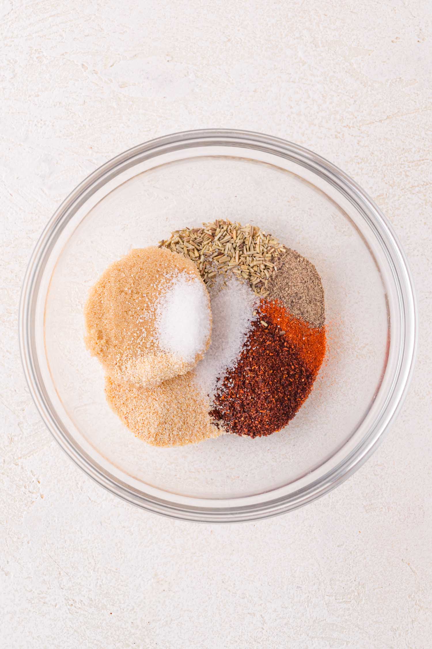 A glass bowl containing separate piles of various ground spices and seasonings, including salt, pepper, paprika, cumin, and garlic powder, on a light surface.