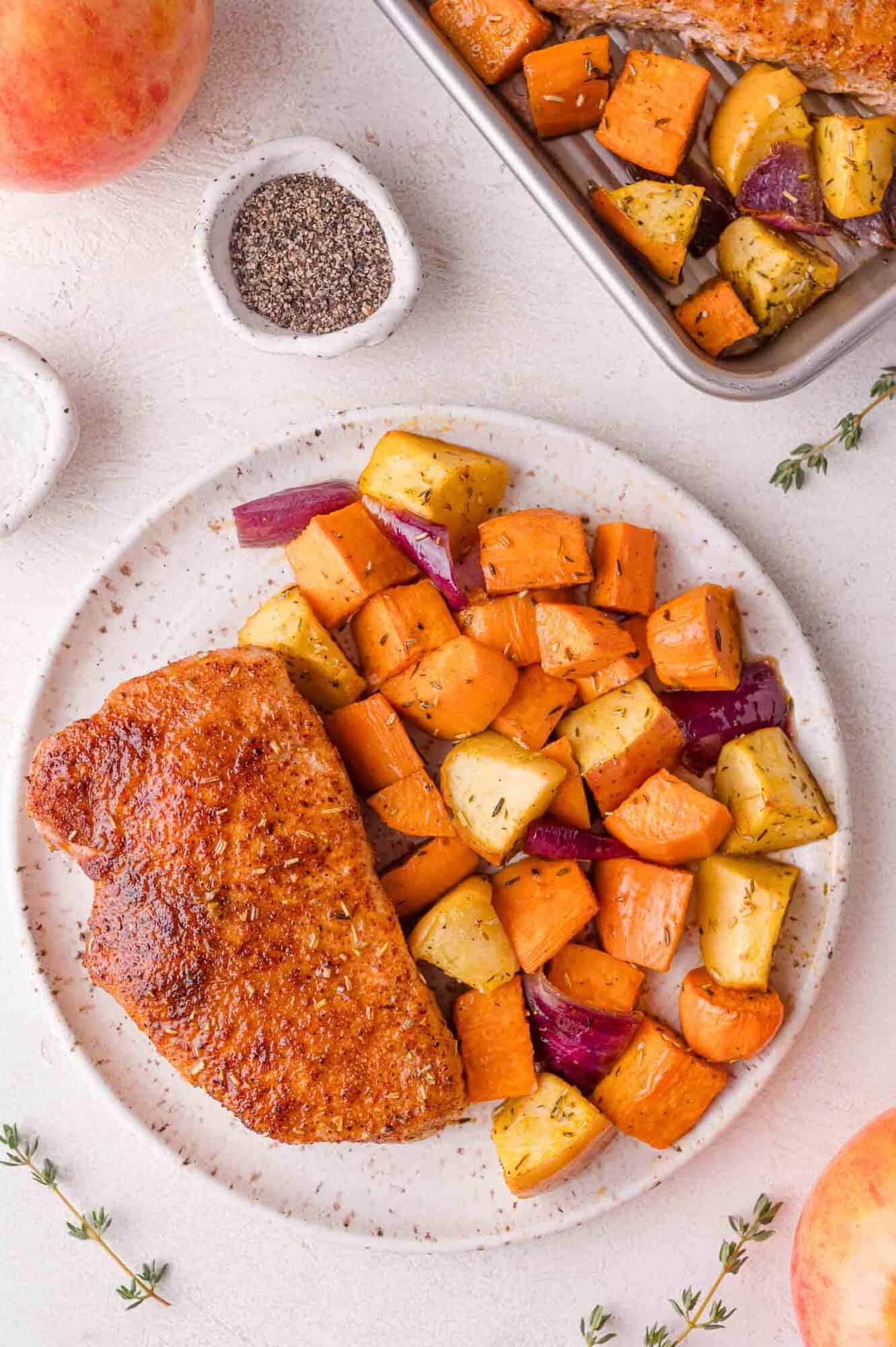 A plate with a seasoned pork chop and roasted vegetables, including sweet potatoes, apples, and red onions, sits on a white surface next to a pan of more vegetables.