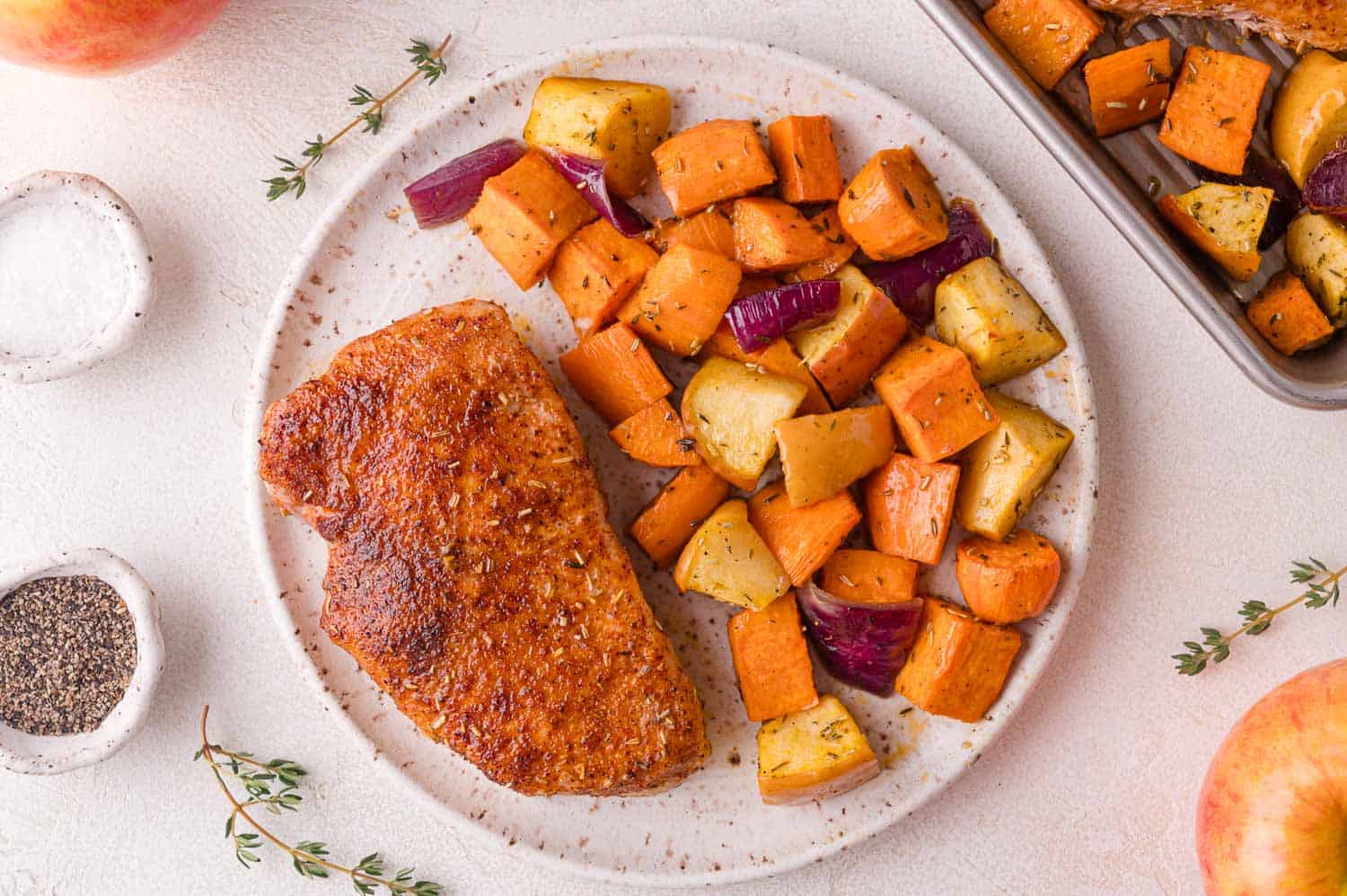A plate with a seasoned cooked pork chop and roasted sweet potatoes, red onions, and apples. Salt, pepper, and thyme garnish are nearby.