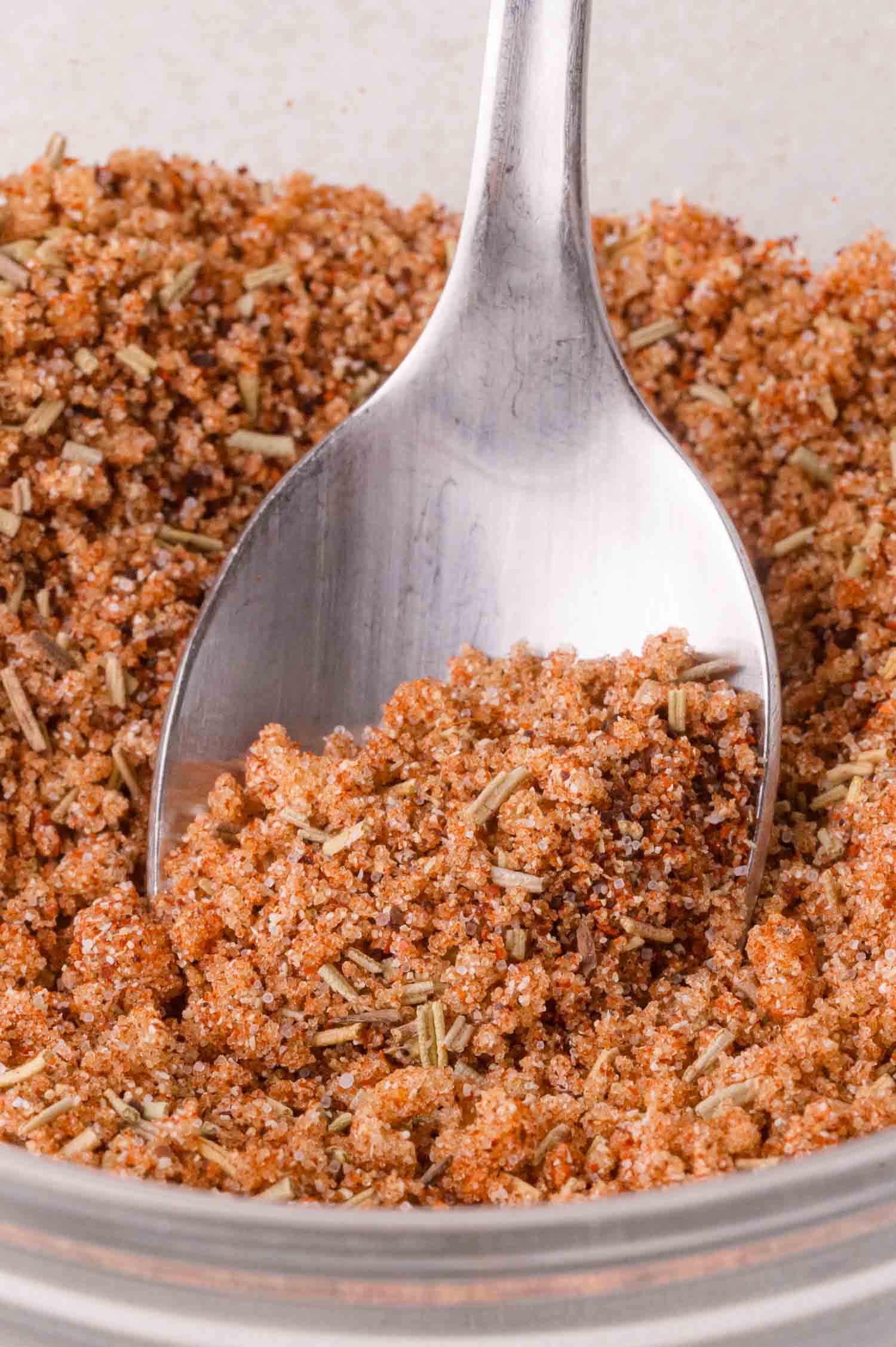 A close-up of a spoon scooping a mixture of spices and herbs from a glass container.