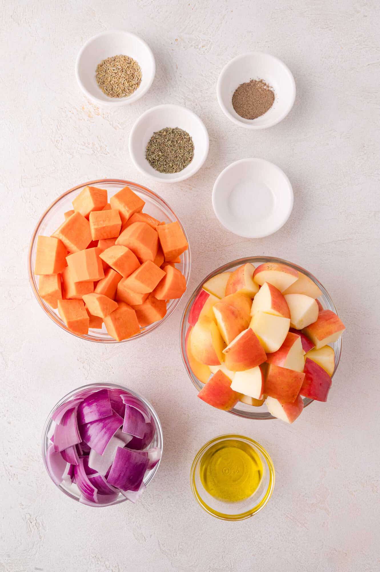 Bowls of diced sweet potatoes, chopped apples, red onion, olive oil, and small dishes of herbs and seasonings arranged on a light surface.