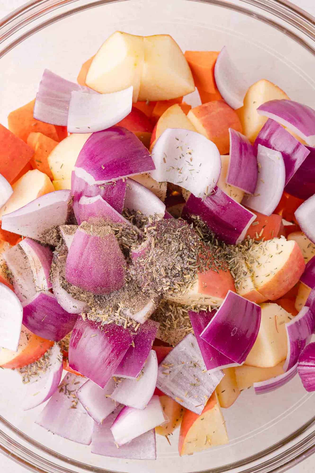 Chopped red onions, apples, and sweet potatoes in a glass bowl, topped with dried herbs and ground black pepper, ready for mixing.