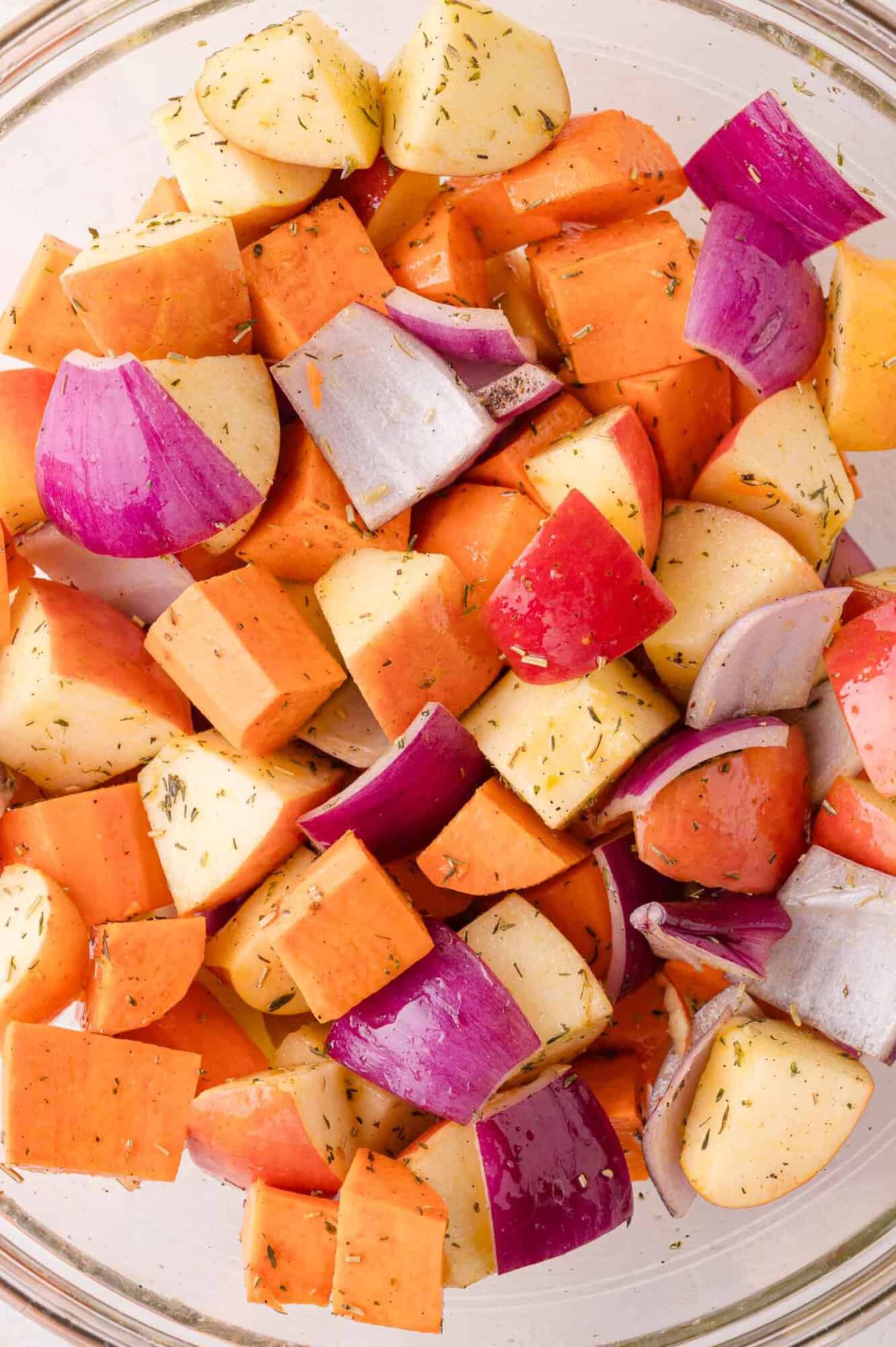 Chopped sweet potatoes, apples, and red onions seasoned with herbs in a glass bowl.
