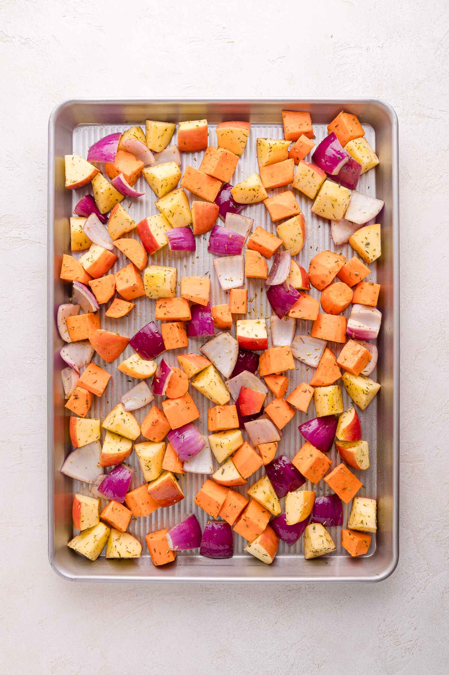 A sheet pan with chopped sweet potatoes, red onions, and apples seasoned with herbs, ready for roasting.