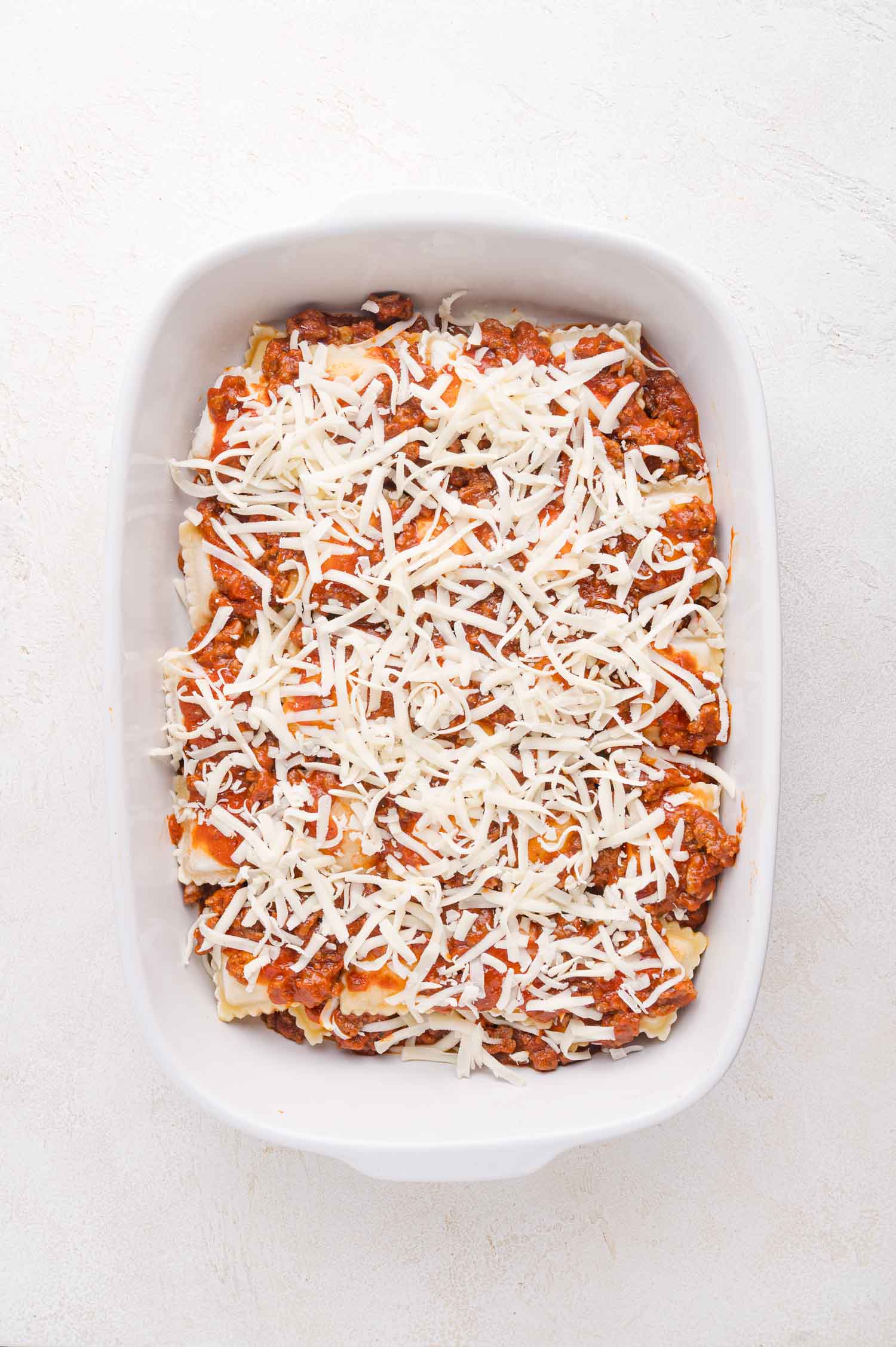 A white baking dish filled with layered baked ravioli, meat sauce, and shredded cheese, ready to be baked.