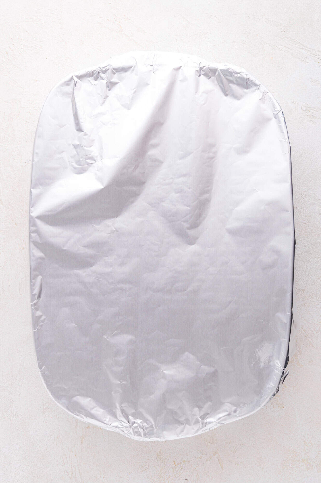 A rectangular baking dish filled with baked ravioli is covered tightly with a sheet of aluminum foil, placed on a light-colored surface.