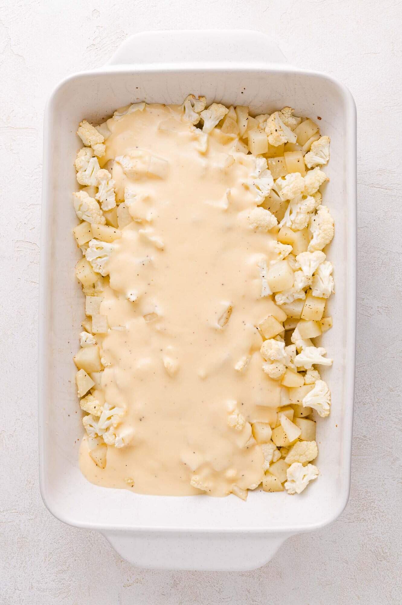 A white baking dish filled with chopped cauliflower and potatoes, partially covered with a layer of creamy cheese sauce.