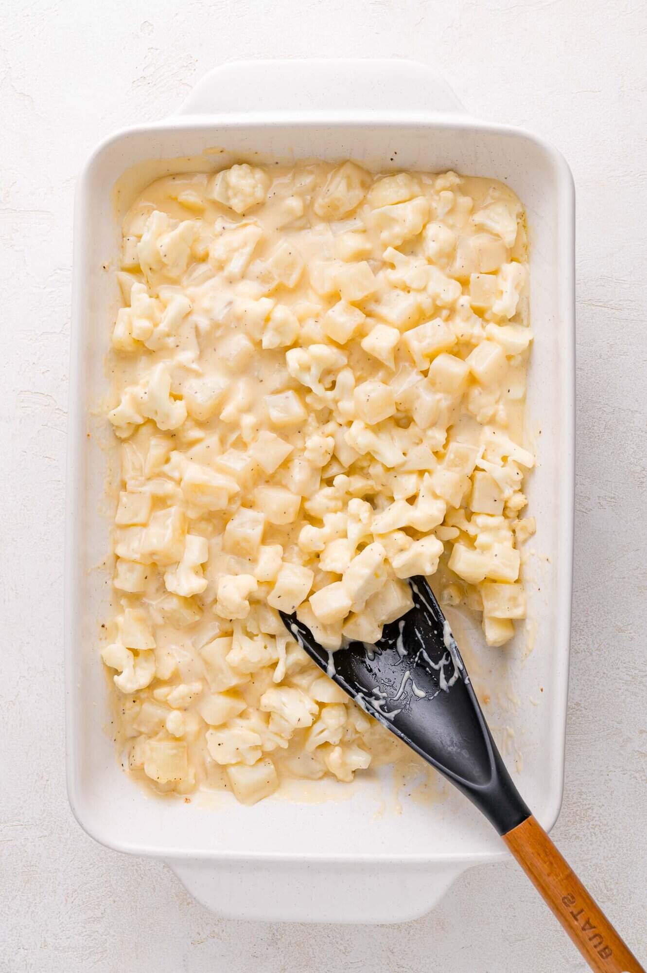 A baking dish filled with diced potatoes mixed in a creamy cheese sauce, with a black spatula resting inside.