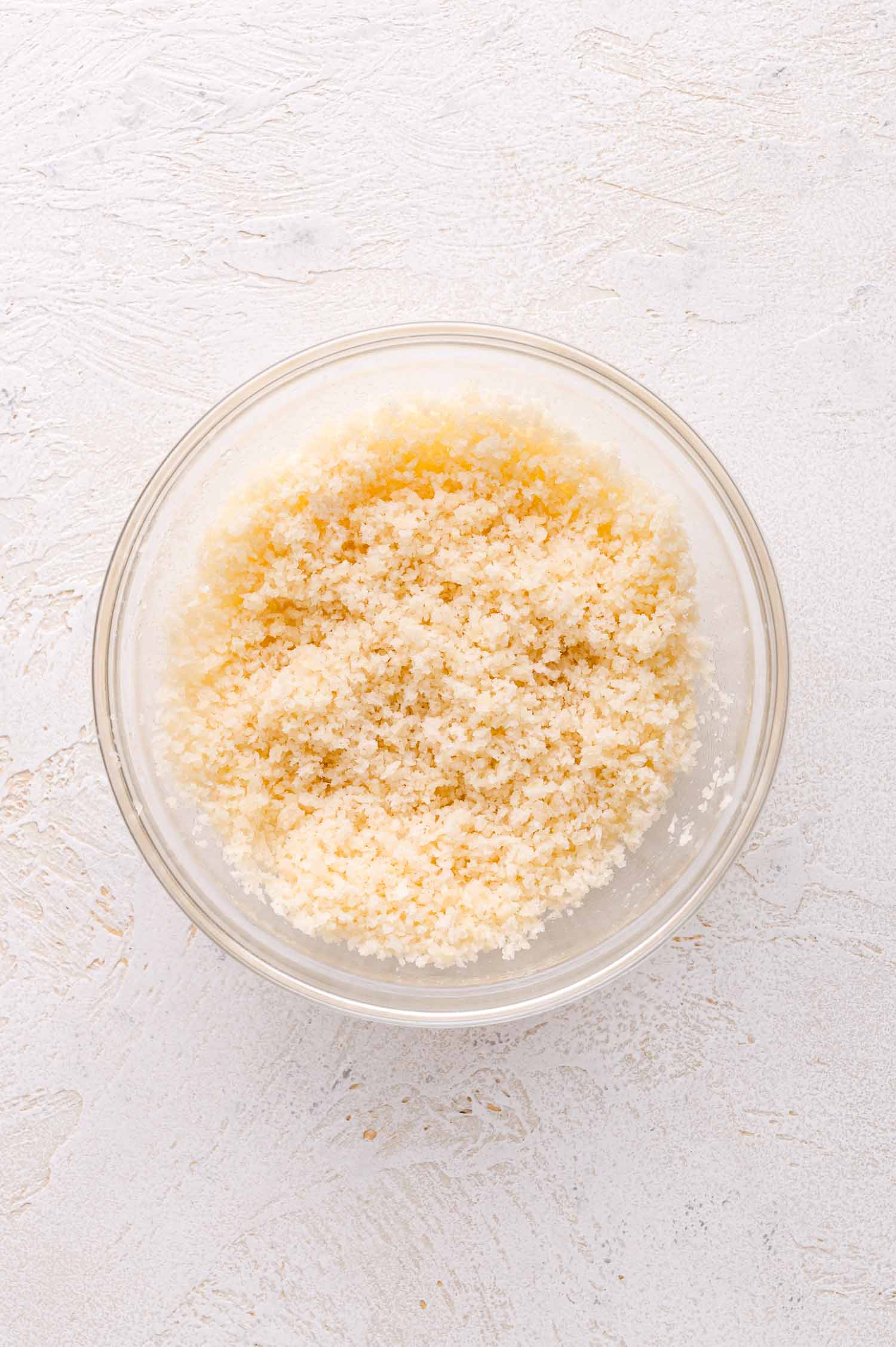 A clear glass bowl filled with light-colored panko breadcrumbs sits on a textured white surface.