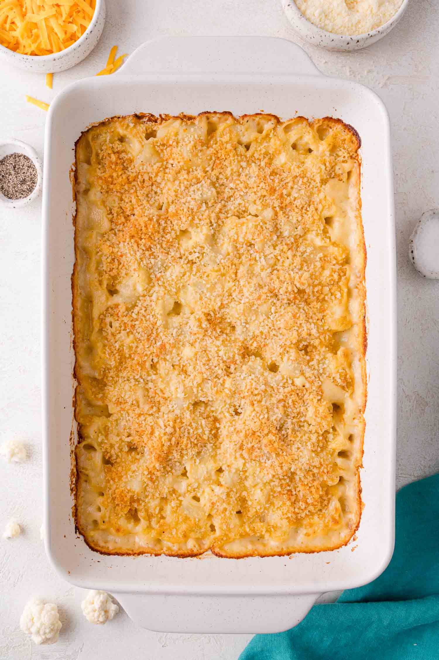 Baked cauliflower and potato casserole with a golden breadcrumb topping in a white rectangular dish, surrounded by bowls of shredded cheese and pepper.