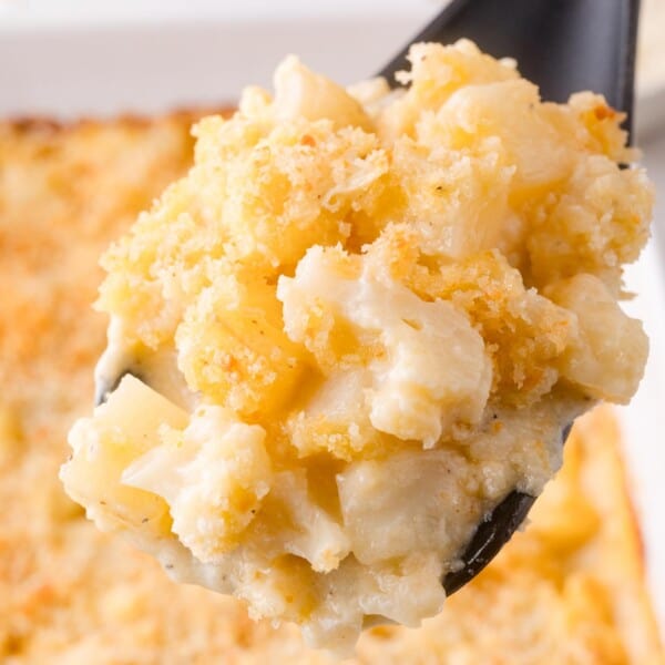A close-up of a serving spoon holding a portion of cheesy, baked cauliflower and potato casserole with a golden breadcrumb topping.