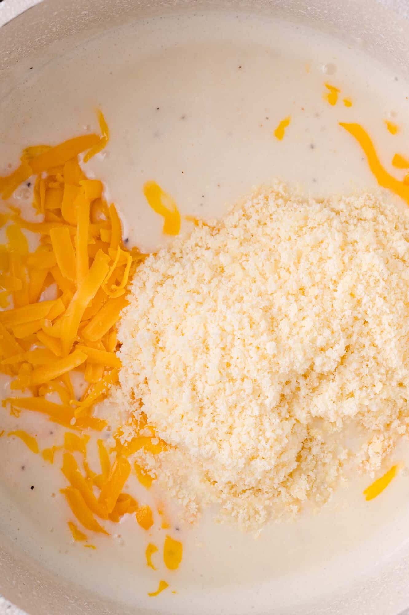 Shredded cheddar cheese and grated parmesan cheese added to a creamy white sauce in a pot.