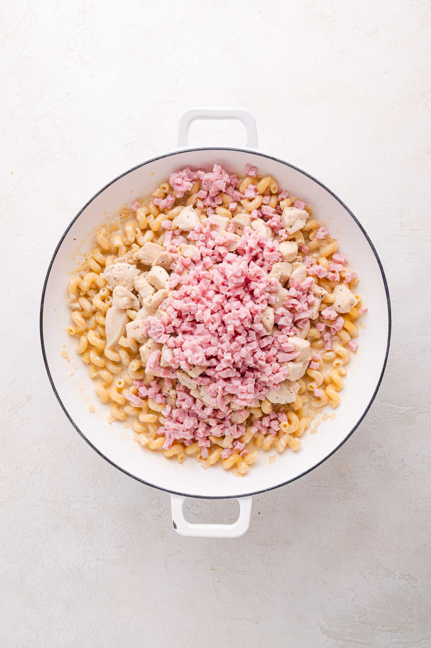 A white pot contains cooked pasta, diced cooked chicken, and chopped ham on a light surface.