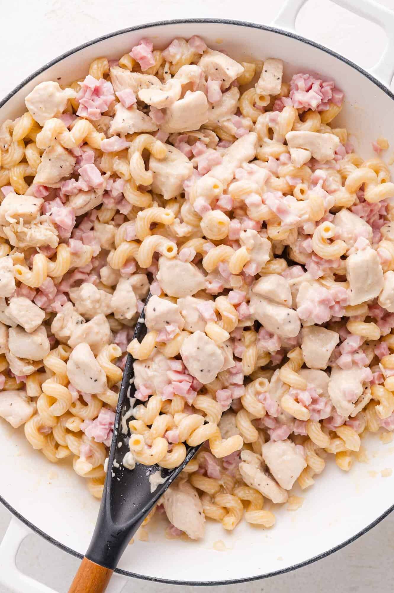 A skillet filled with cooked cavatappi pasta, diced chicken, and chopped ham coated in a creamy white sauce, with a black serving spoon.