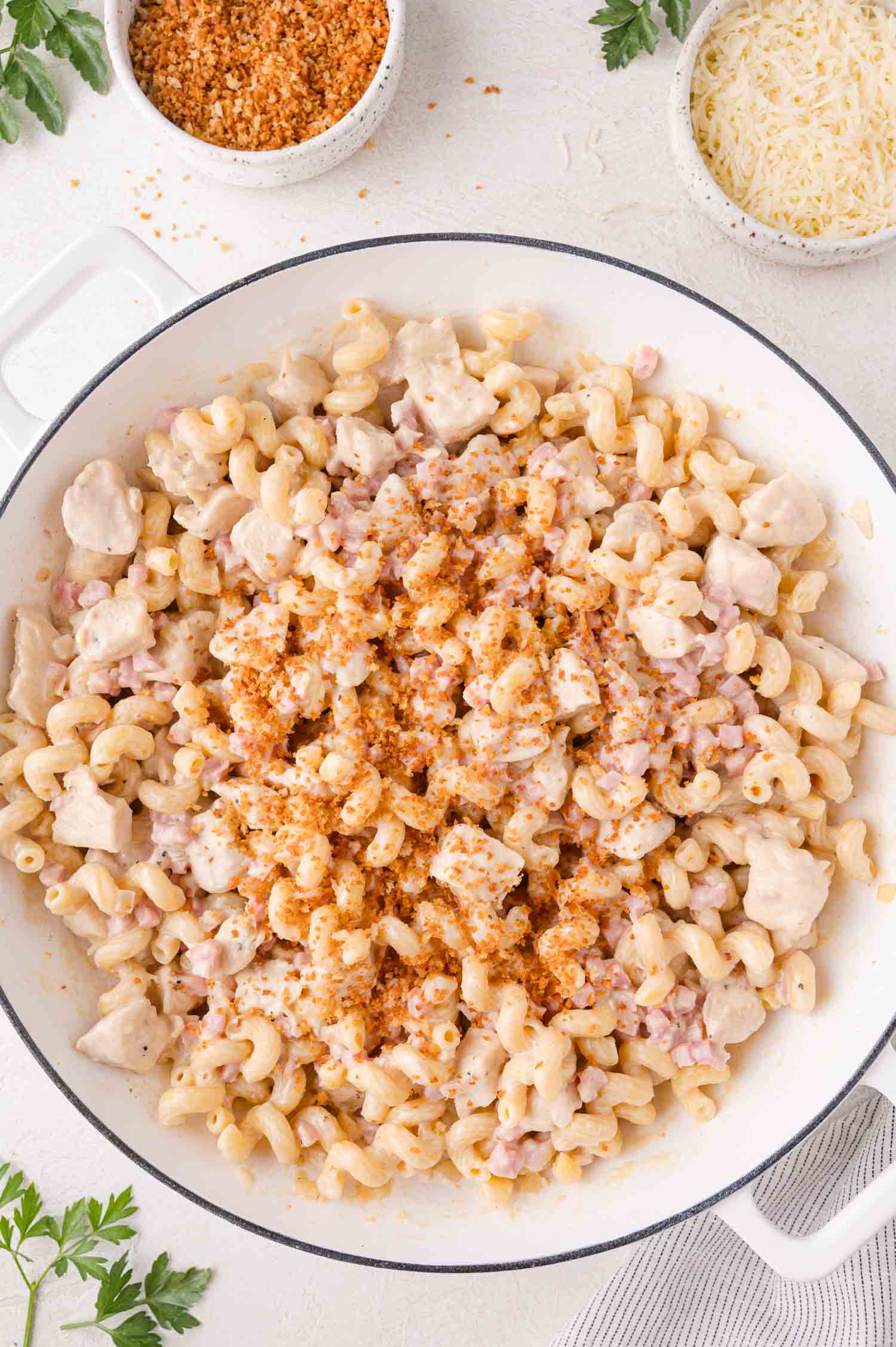 A white skillet filled with creamy pasta, diced chicken, and ham, topped with toasted breadcrumbs. Bowls of grated cheese and breadcrumbs are nearby on a light surface.