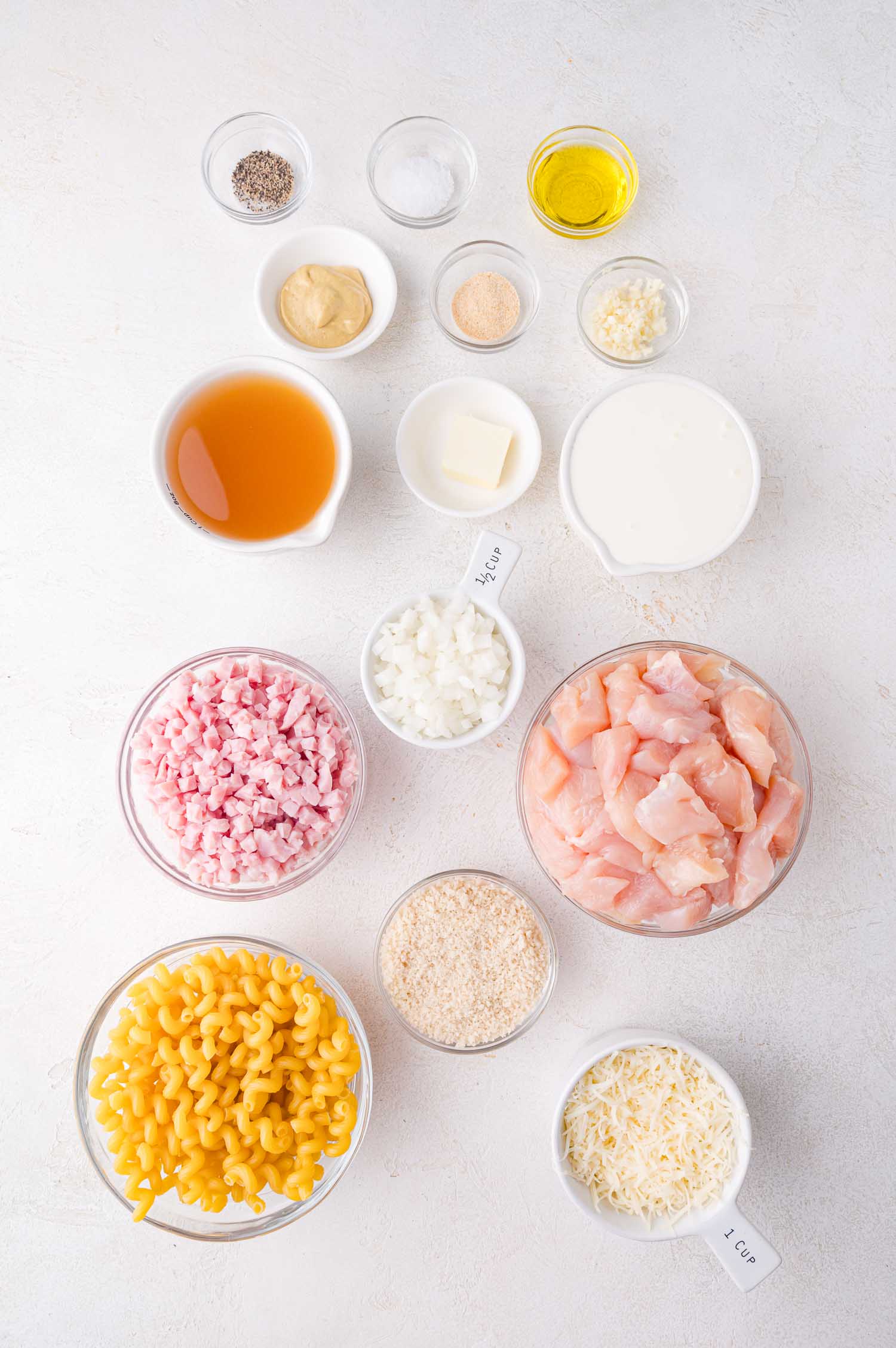 Ingredients for a pasta dish arranged on a white surface, including diced chicken, pasta, diced ham, chopped onions, broth, cream, cheese, breadcrumbs, and seasonings in bowls.