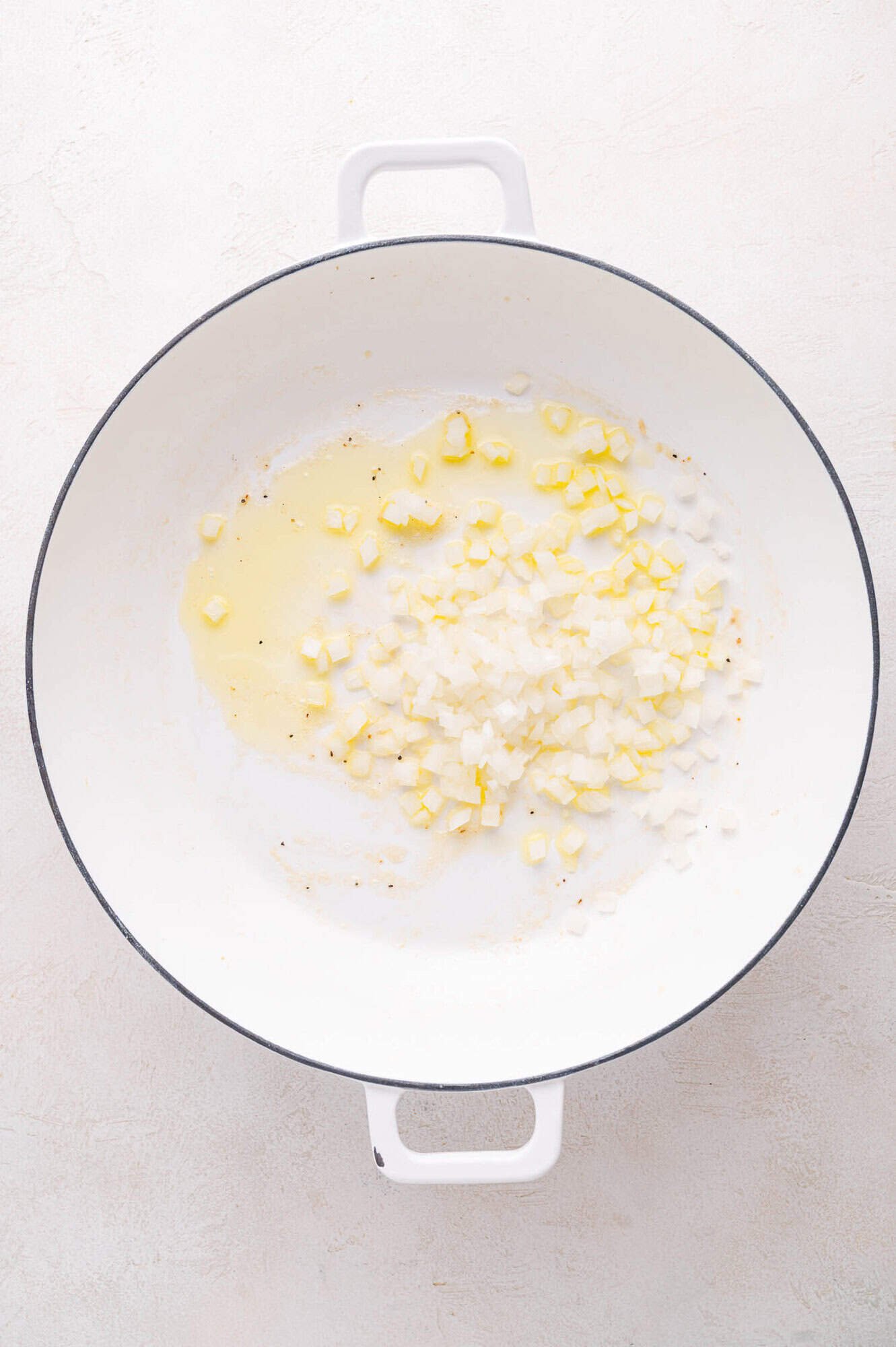 Diced onions sautéing in oil in a white enameled pan on a light surface.