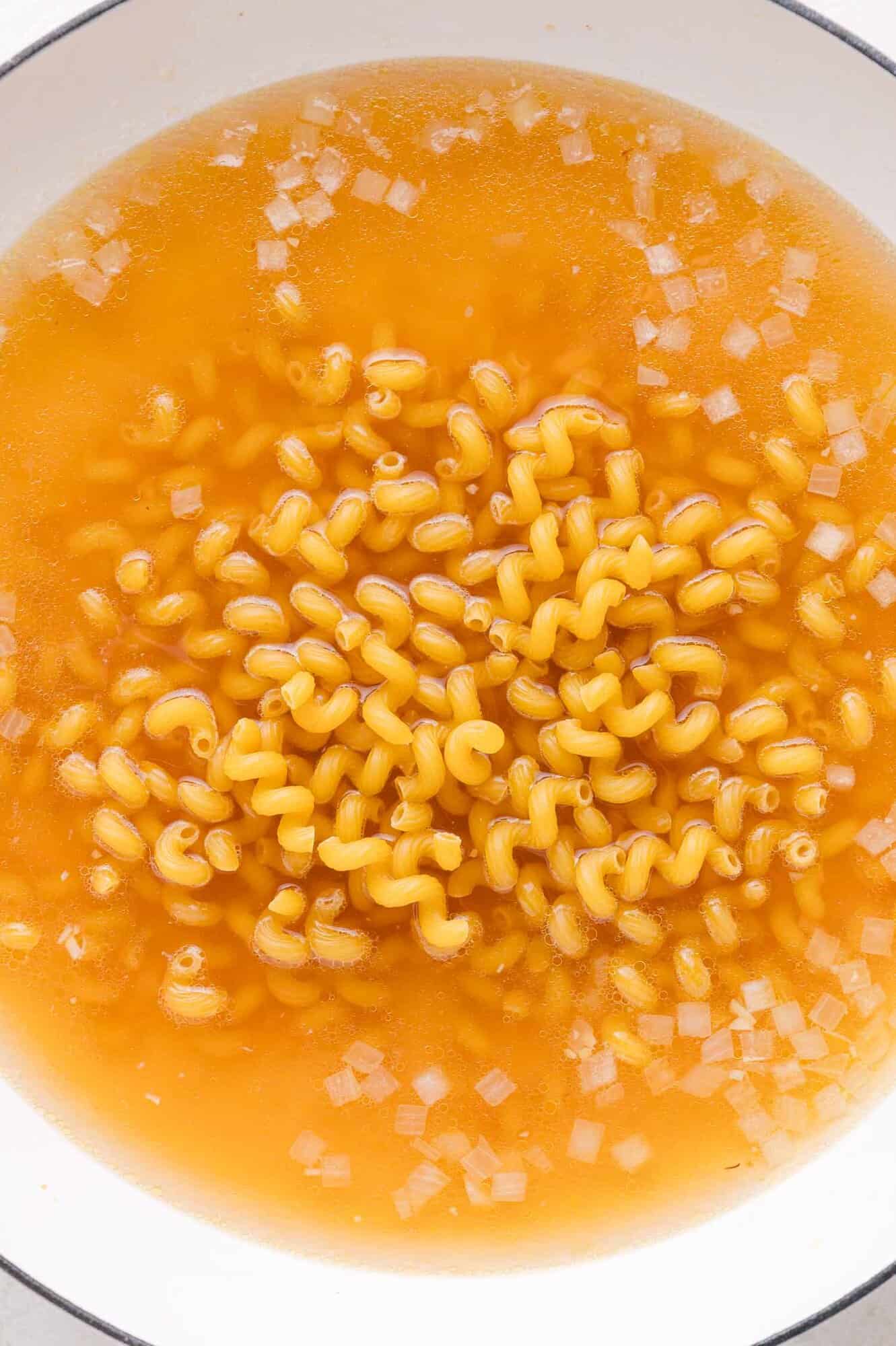 A pot of uncooked curly pasta, diced onions, and broth.