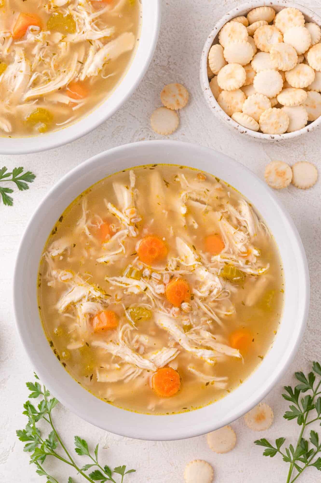 Two bowls of chicken farro soup with carrots and celery, next to a small bowl of round oyster crackers and parsley garnish on a white surface.