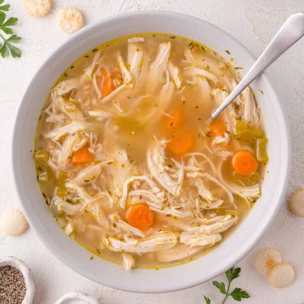 A bowl of chicken soup with farro, shredded chicken, carrots, and celery, served with a spoon. Oyster crackers and parsley are scattered on the white surface nearby.
