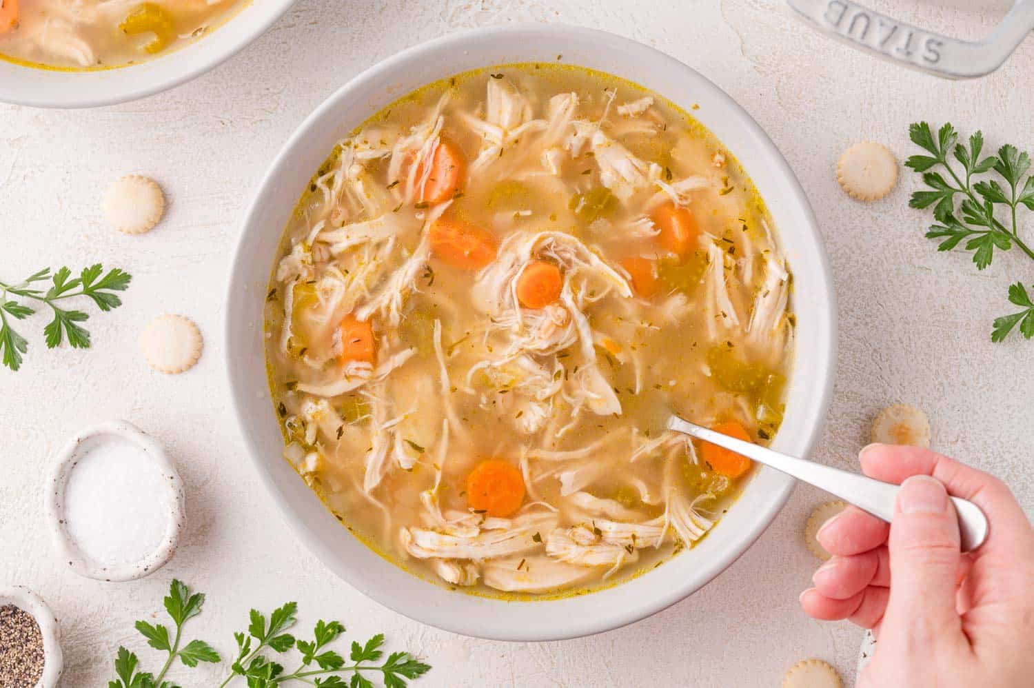 A bowl of chicken soup with shredded chicken, carrots, and celery, surrounded by parsley, salt, pepper, and oyster crackers; a hand holds a spoon in the soup.