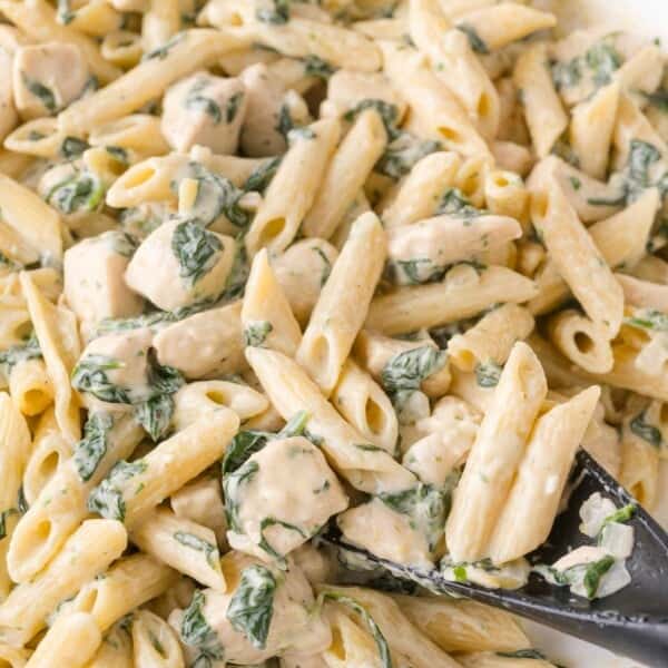 Close-up of creamy penne pasta mixed with pieces of chicken and spinach, being served with a black spoon.