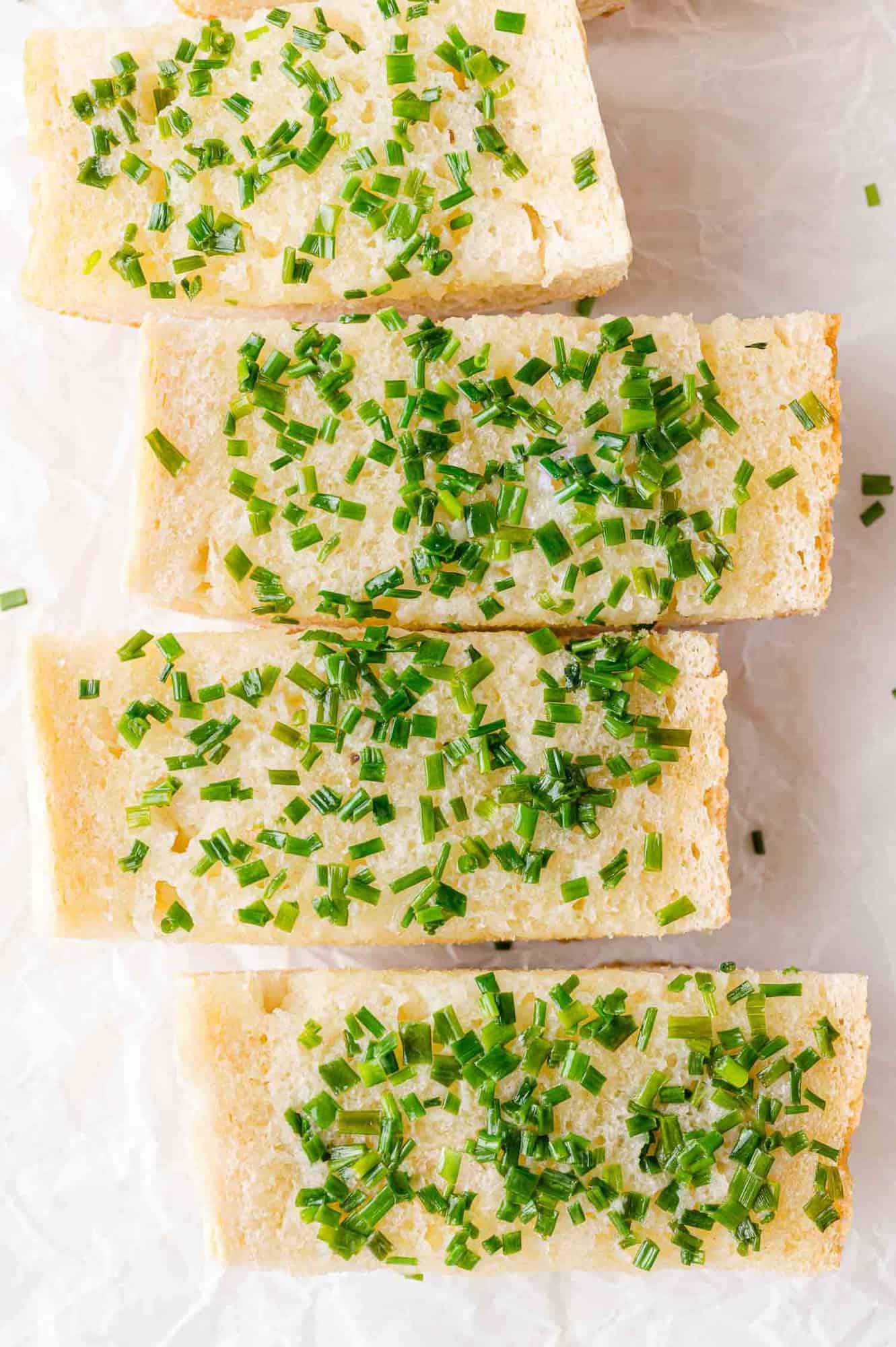 Four rectangular slices of bread topped with chopped fresh chives are arranged in a row on a white surface.