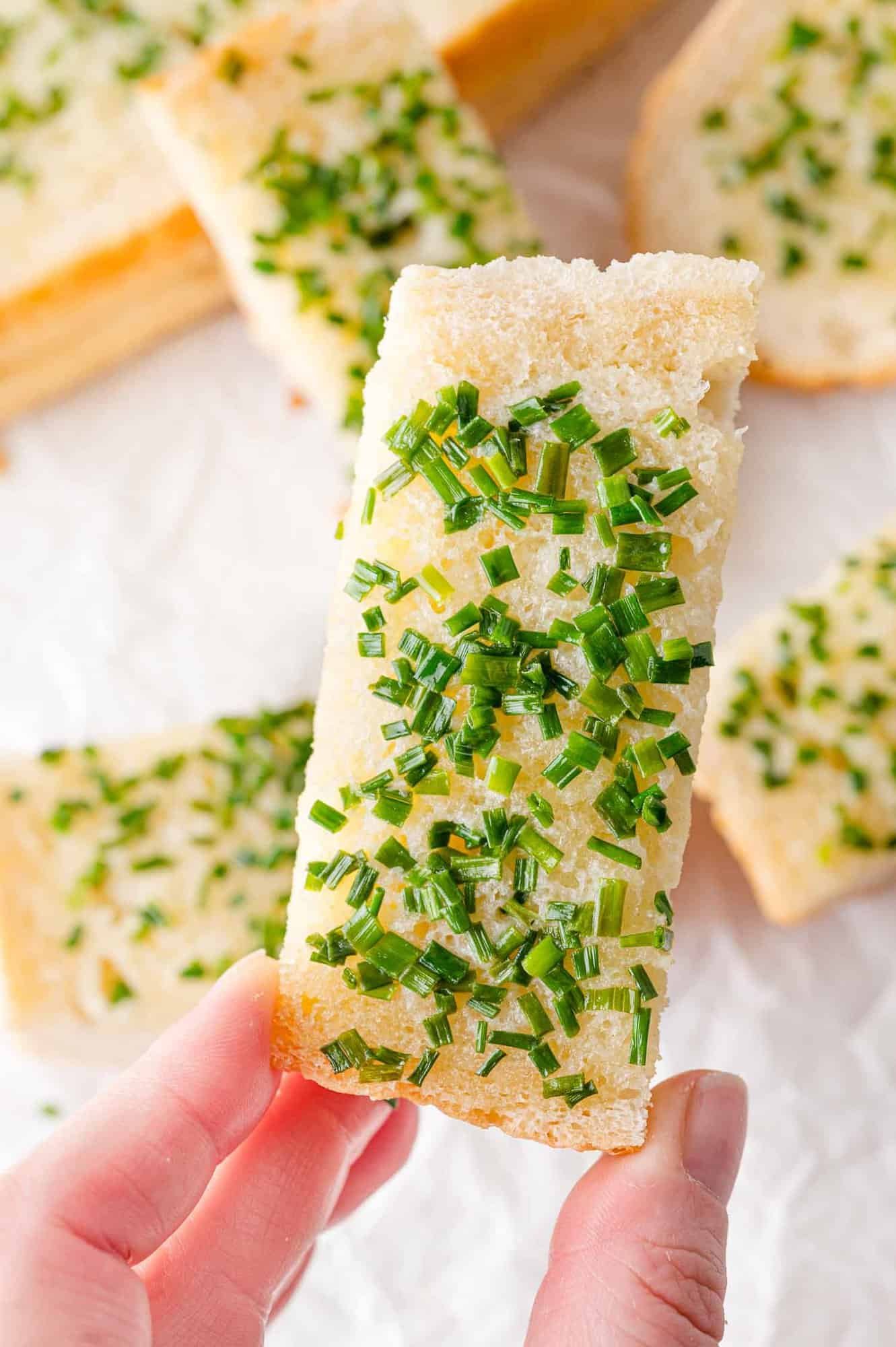 A hand holding a rectangular slice of bread topped with chopped chives, with more similar slices in the background.