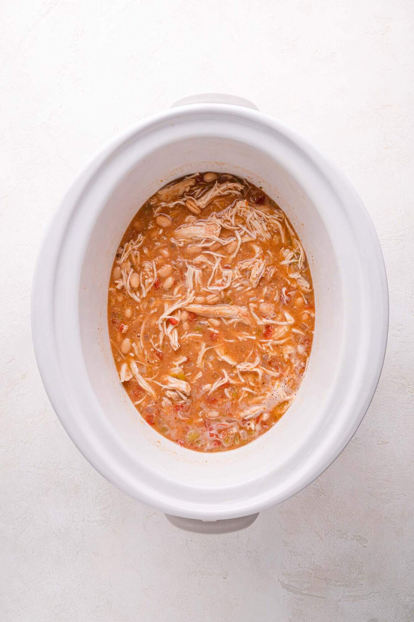 A white slow cooker filled with shredded chicken in a broth with beans, tomatoes, and visible seasonings.