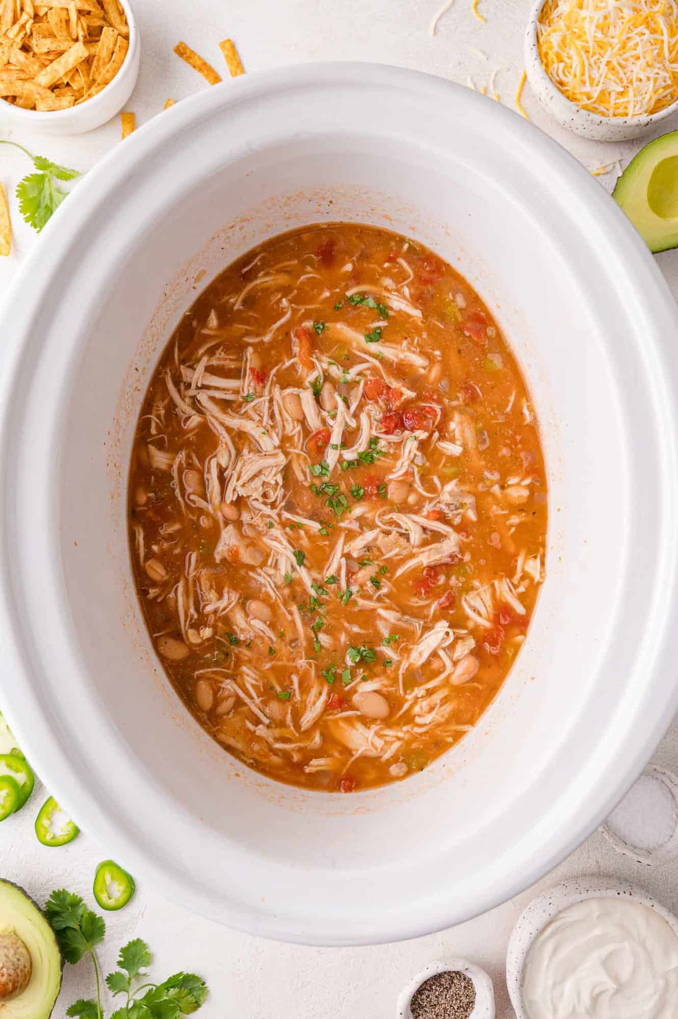 Overhead view of shredded chicken chili in a white slow cooker, surrounded by bowls of toppings like avocado, cheese, sour cream, jalapeño, and tortilla strips.