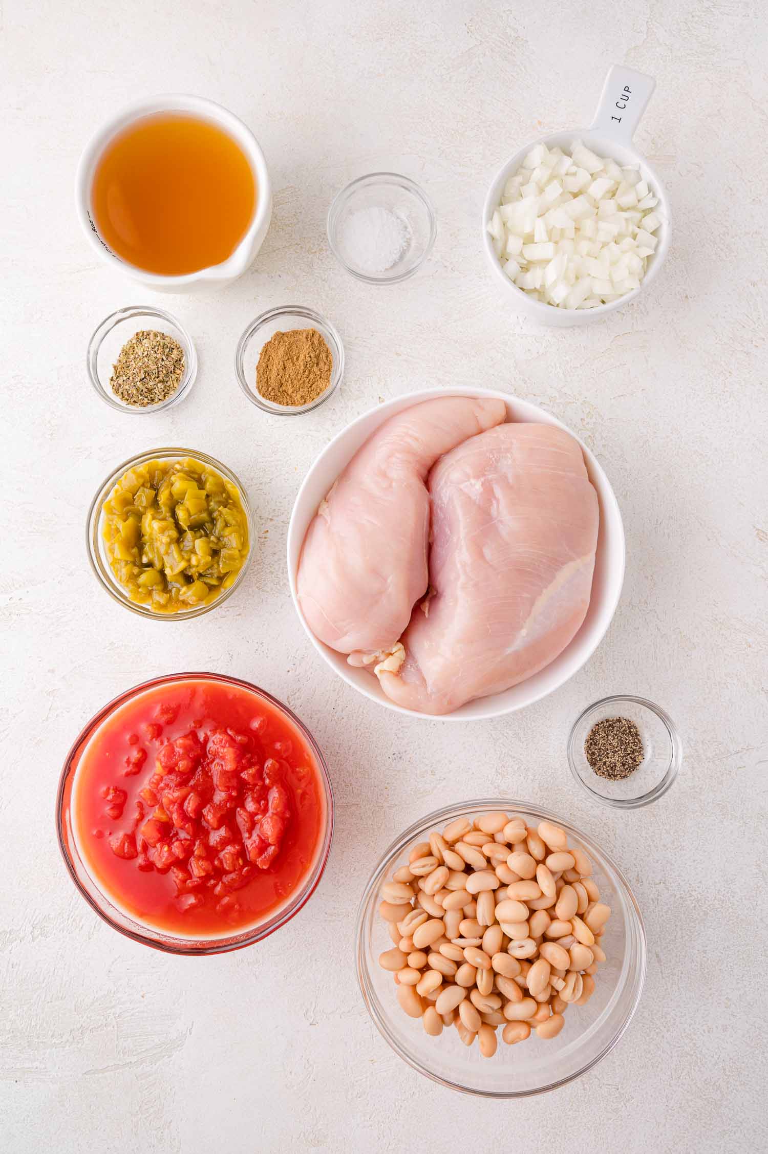 Overhead view of raw chicken breasts, diced tomatoes, white beans, chopped onions, diced green chiles, chicken broth, and small bowls of spices on a light surface.