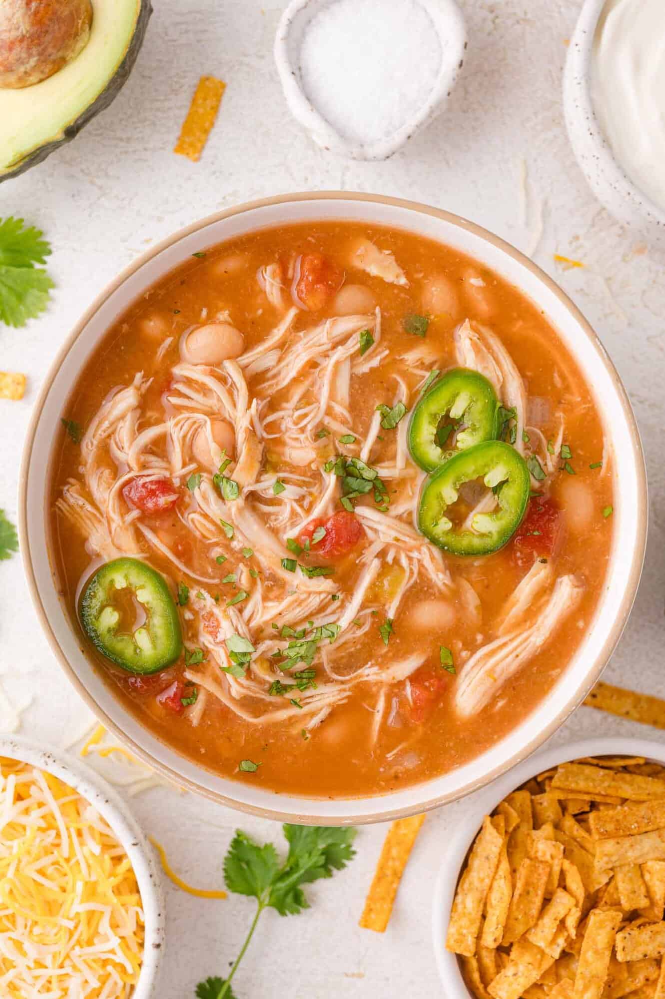 A bowl of crockpot white chicken chili topped with sliced jalapeños and chopped herbs, surrounded by shredded cheese, tortilla strips, an avocado, and a small bowl of salt.