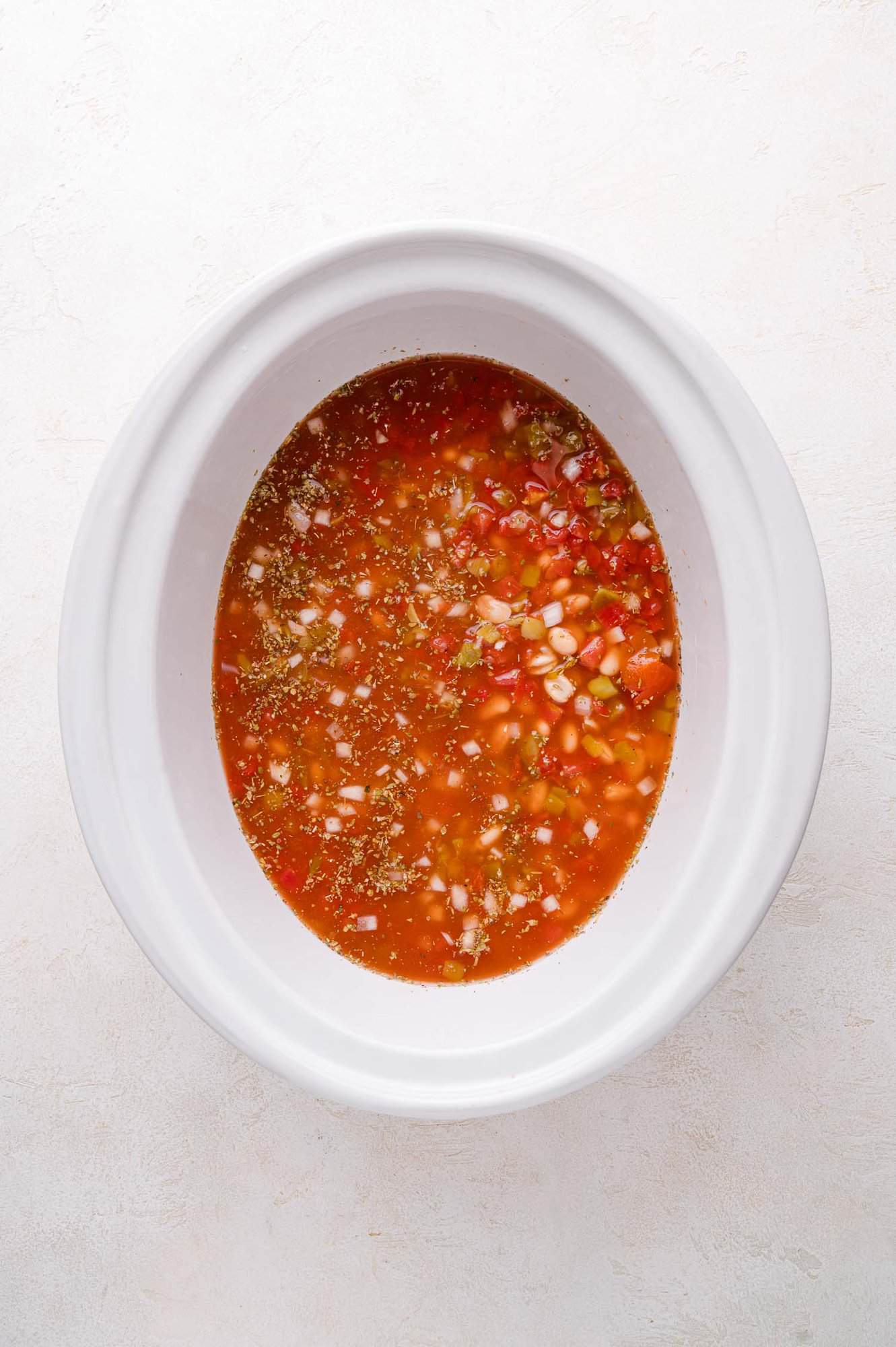 An overhead view of a white slow cooker filled with a tomato-based mixture containing diced onions, peppers, and seasonings.