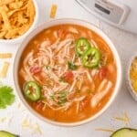 A bowl of chicken chili topped with sliced jalapeños and chopped herbs, next to bowls of shredded cheese and tortilla strips.