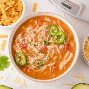 A bowl of chicken chili topped with sliced jalapeños and chopped herbs, next to bowls of shredded cheese and tortilla strips.