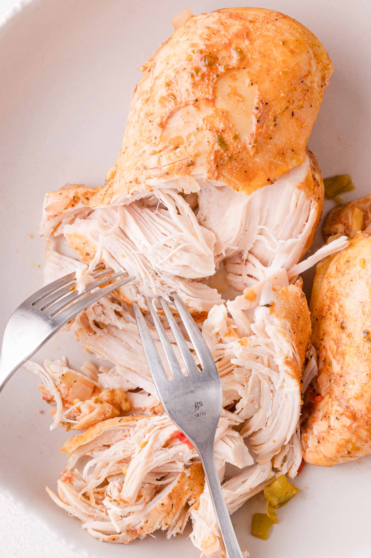 Two forks pulling apart cooked, seasoned chicken breast on a white plate.