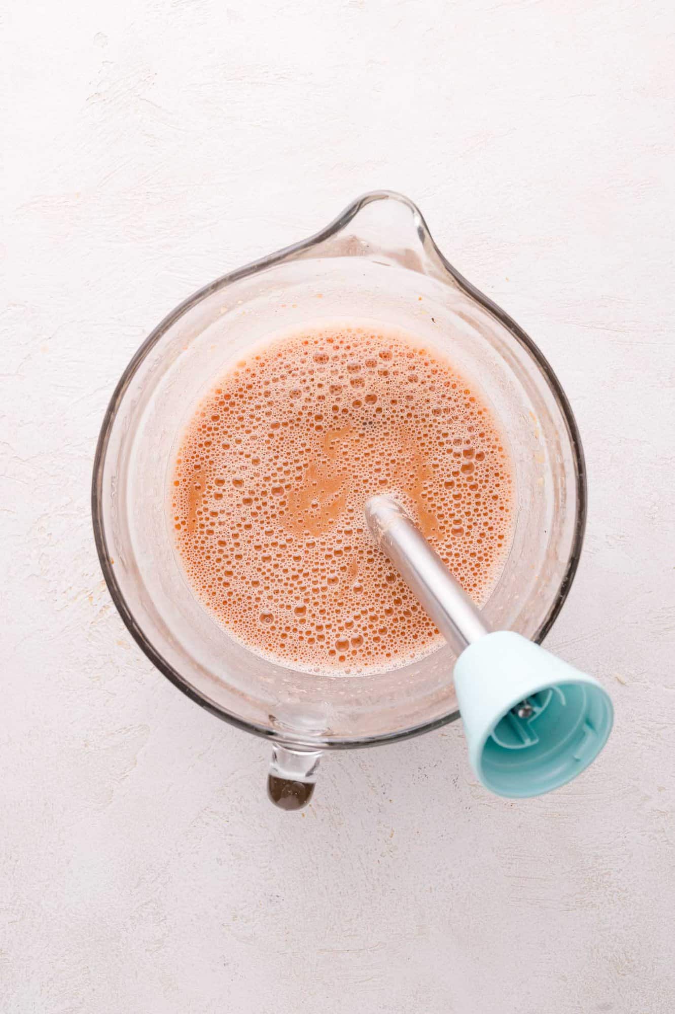 A glass measuring cup filled with a frothy light brown liquid and an immersion blender inside it, viewed from above.