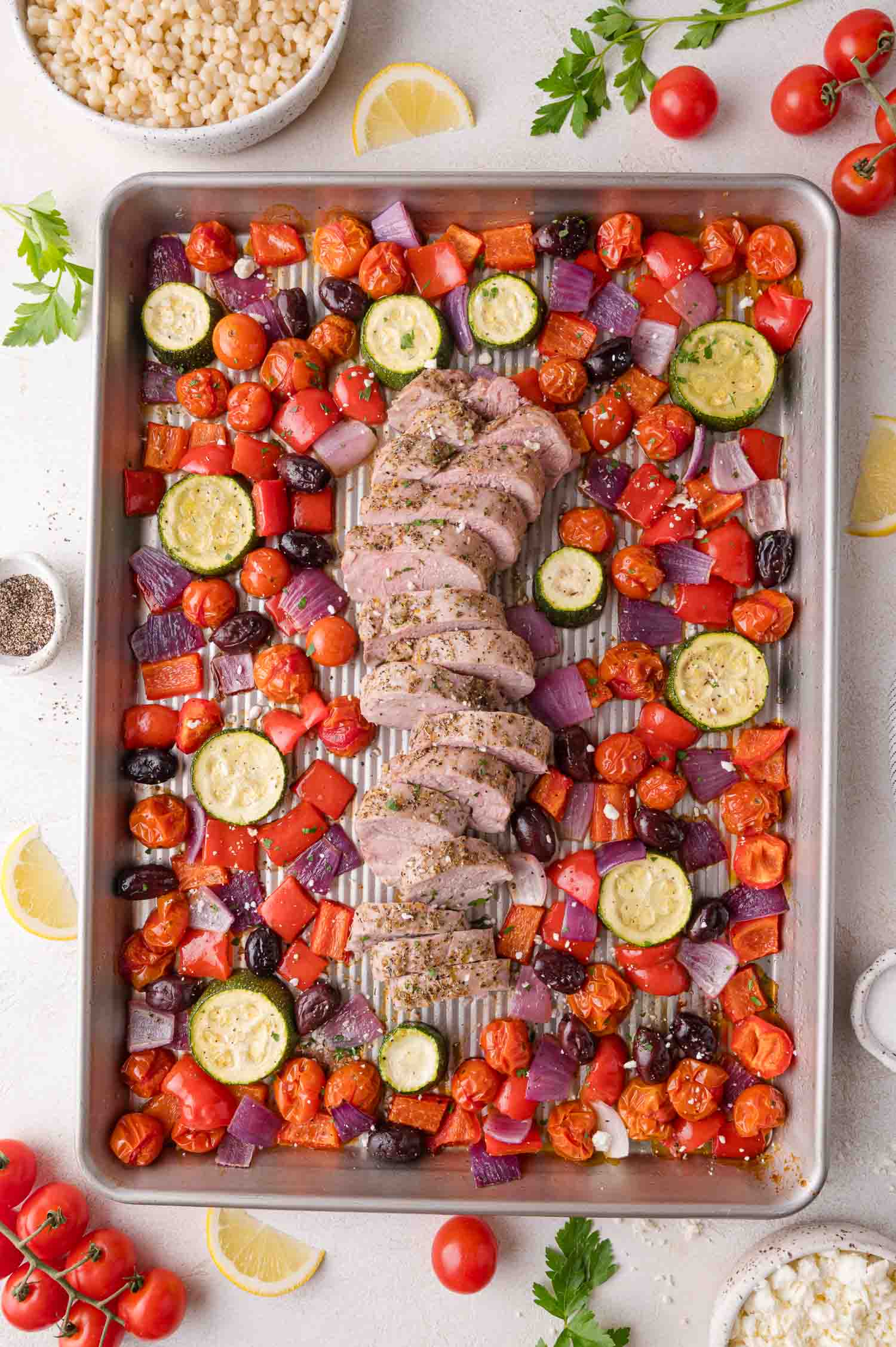 A baking sheet with sliced cooked pork tenderloin surrounded by roasted cherry tomatoes, red onion, zucchini, red bell pepper, and olives.