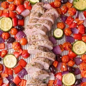 Sliced cooked pork tenderloin on a sheet pan surrounded by roasted cherry tomatoes, zucchini, red bell peppers, red onions, and kalamata olives.