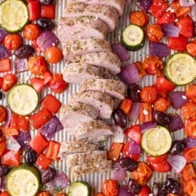 Sliced seasoned pork tenderloin on a baking sheet, surrounded by roasted zucchini, cherry tomatoes, red onion, red bell pepper, and kalamata olives.