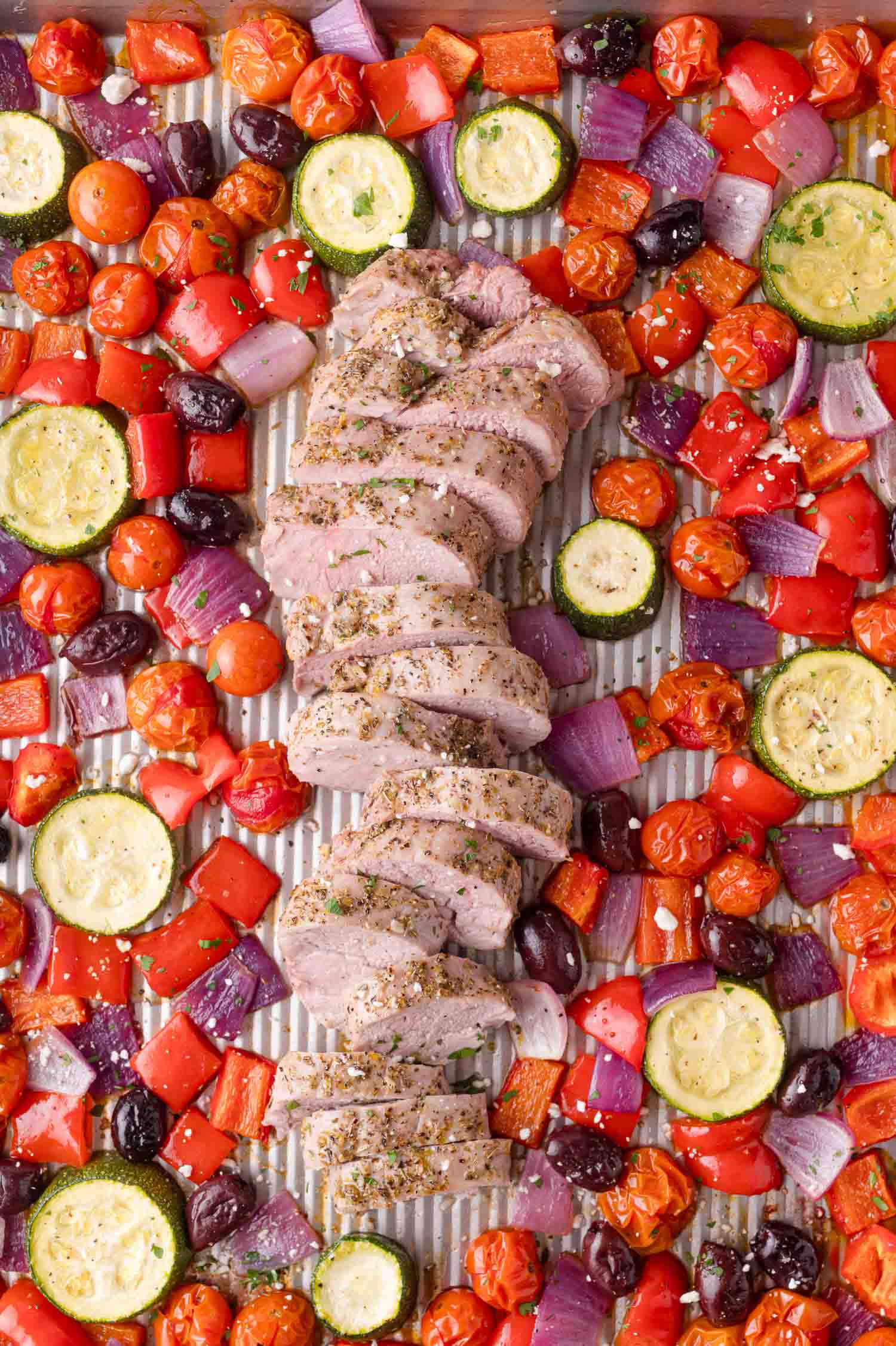 Sliced cooked pork tenderloin on a sheet pan surrounded by roasted cherry tomatoes, zucchini, red bell peppers, red onions, and kalamata olives.