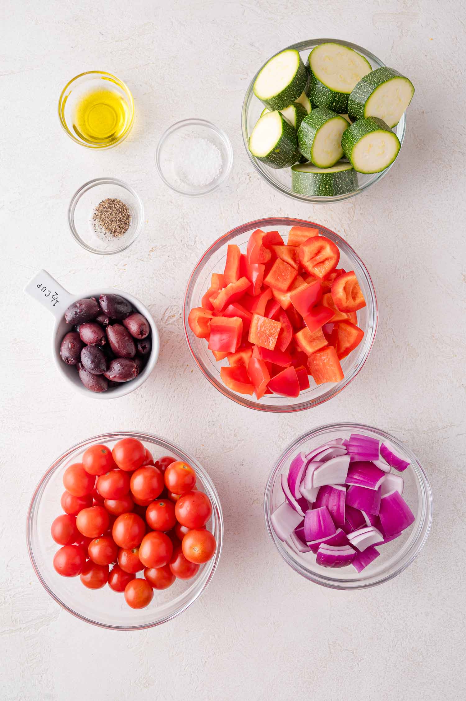 Bowls of chopped zucchini, red bell pepper, red onion, cherry tomatoes, kalamata olives, olive oil, salt, and pepper arranged on a white surface.