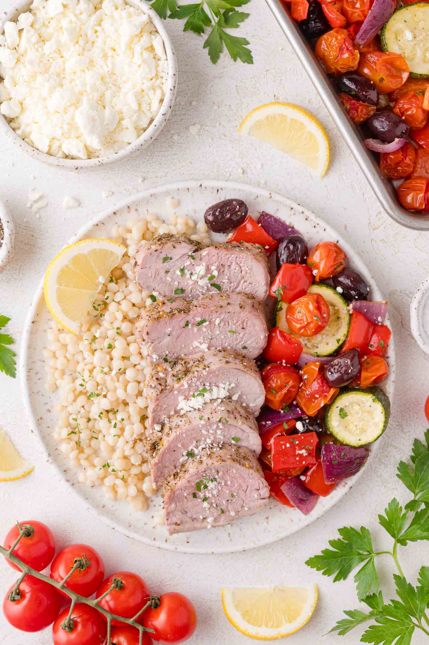 A plate with sliced pork tenderloin, cooked couscous, roasted tomatoes, zucchini, red onion, red bell pepper, and kalamata olives, garnished with herbs and lemon wedges.