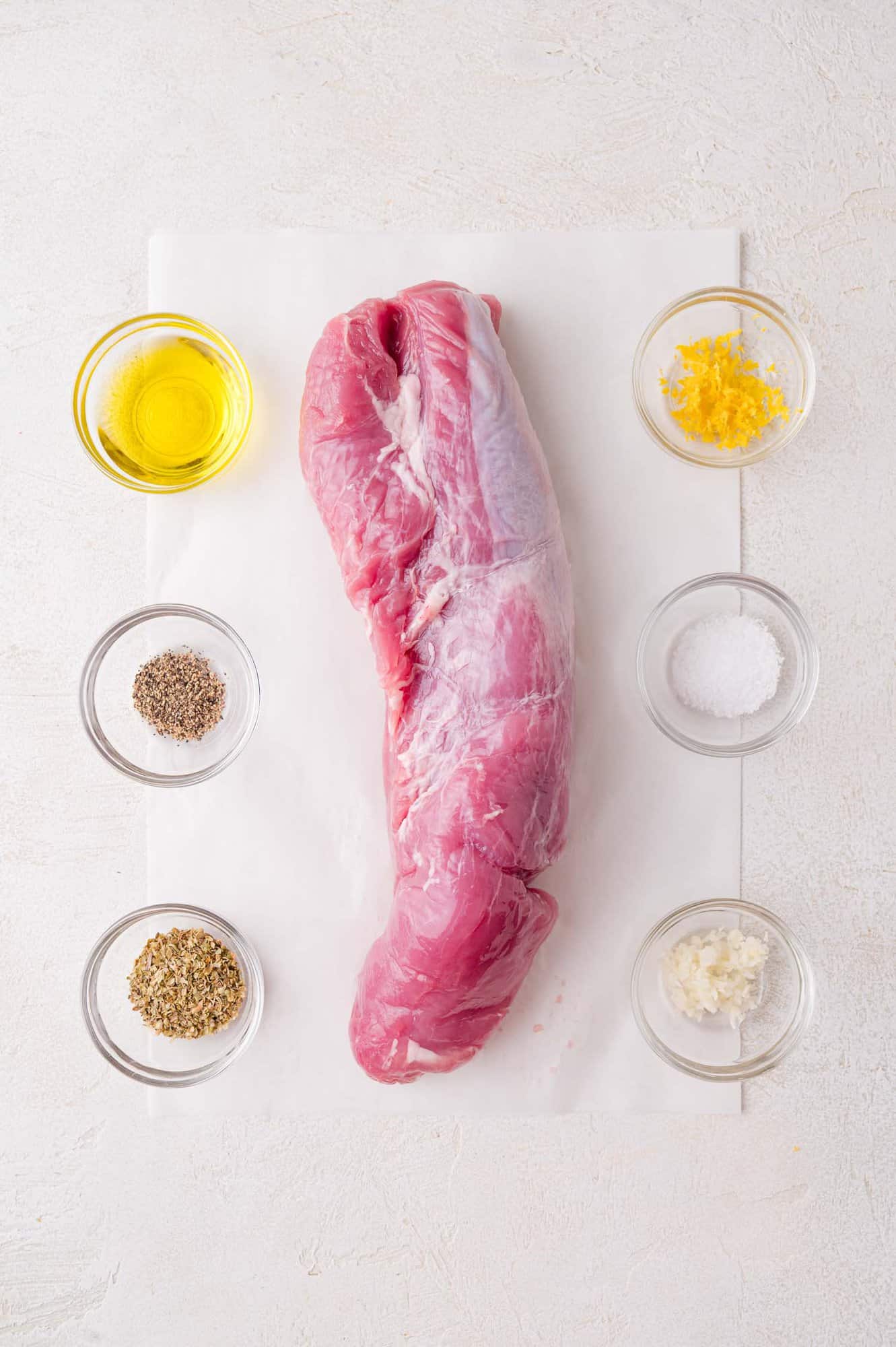 Raw pork tenderloin on parchment paper surrounded by small bowls of olive oil, lemon zest, salt, minced garlic, dried herbs, and ground black pepper.