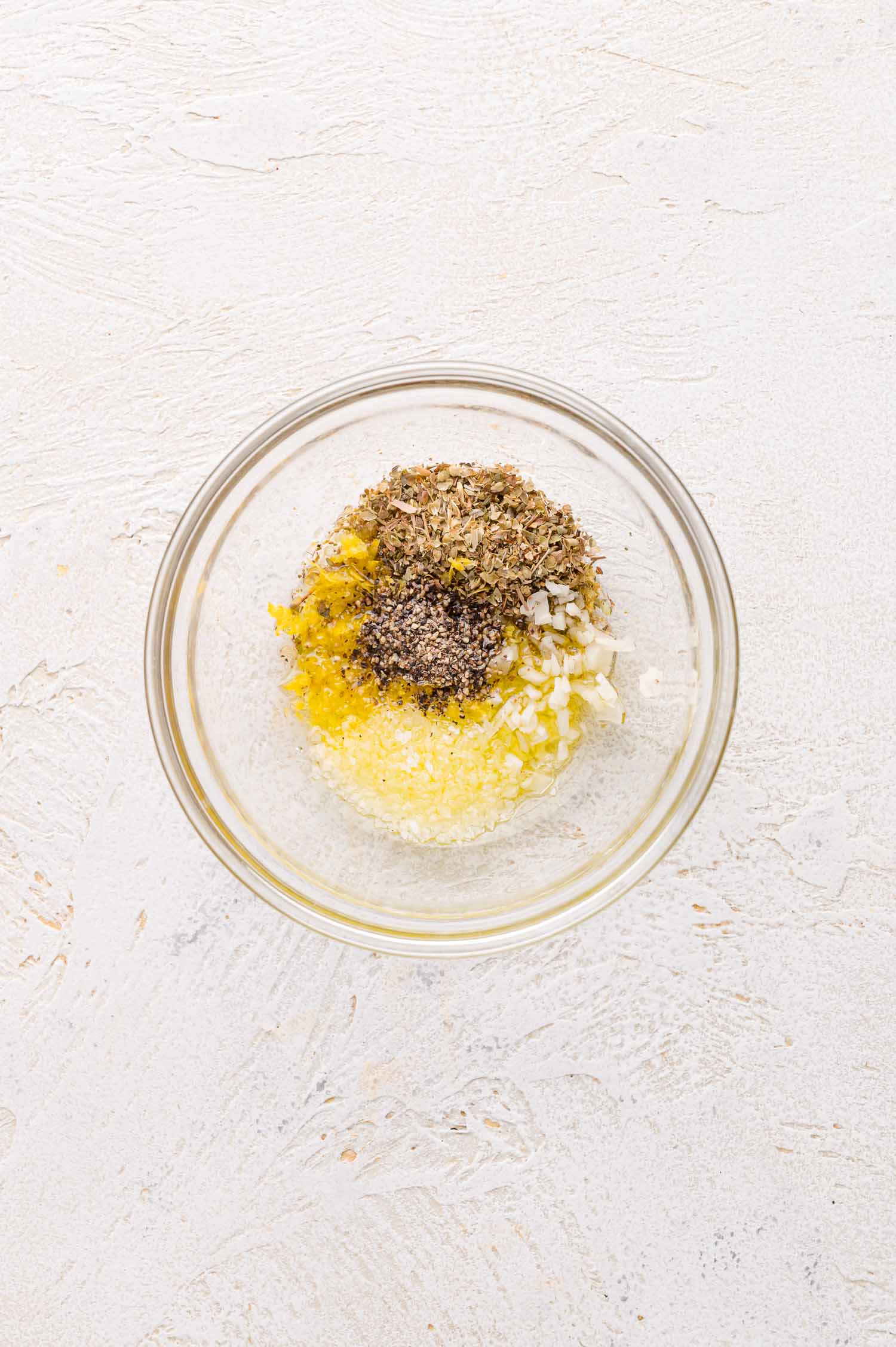 A glass bowl containing minced garlic, dried herbs, black pepper, lemon zest, and olive oil on a textured white surface.