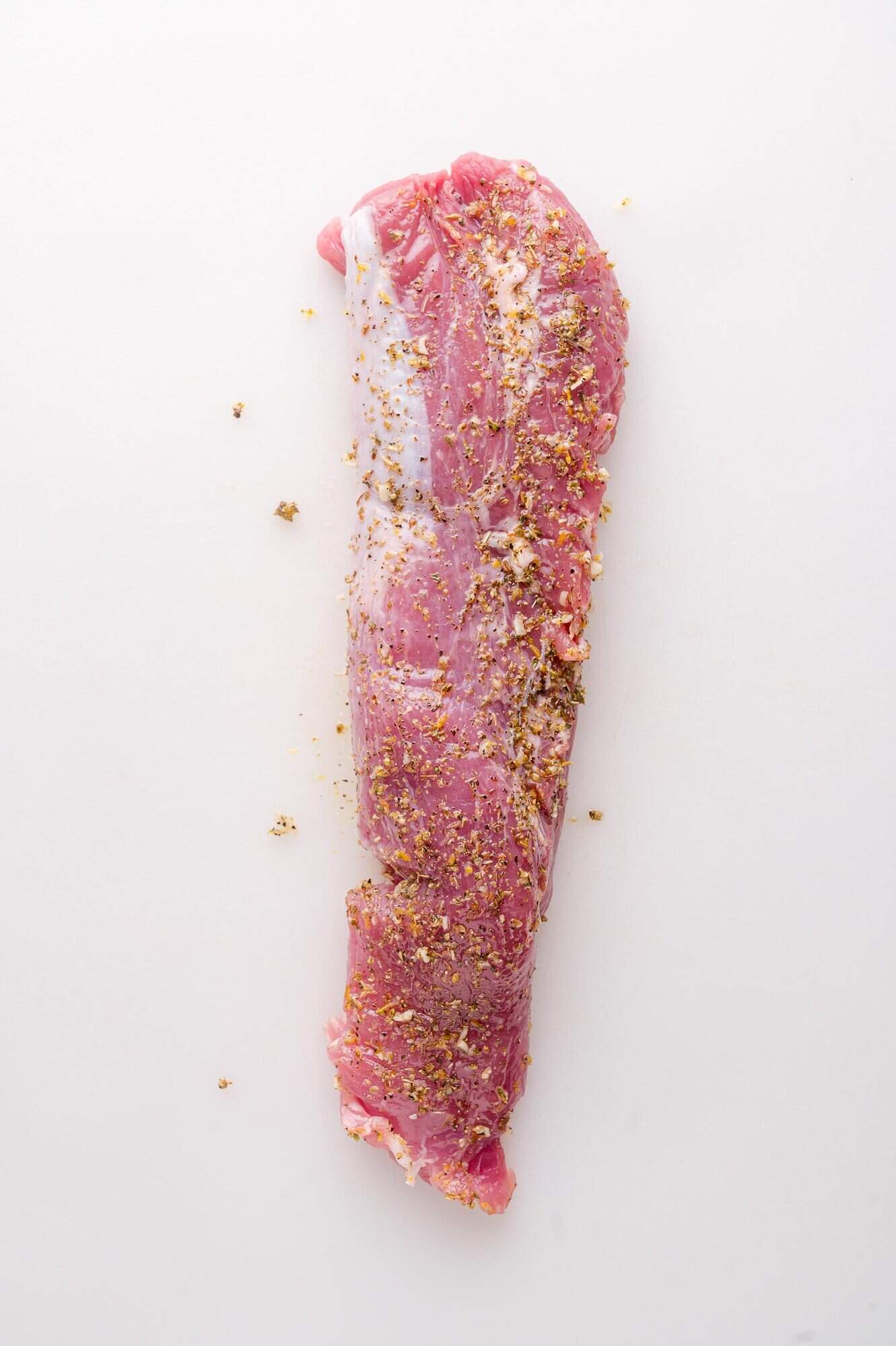 A raw pork tenderloin seasoned with coarse spices is placed on a white surface.