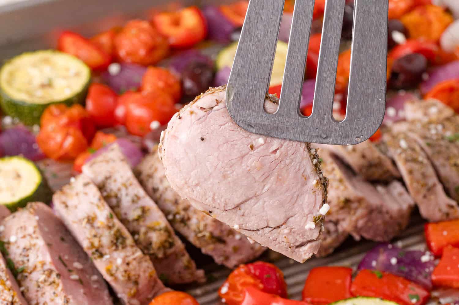 A spatula holds a slice of cooked pork tenderloin above a tray of sliced pork, cherry tomatoes, zucchini, red onion, and red bell pepper.