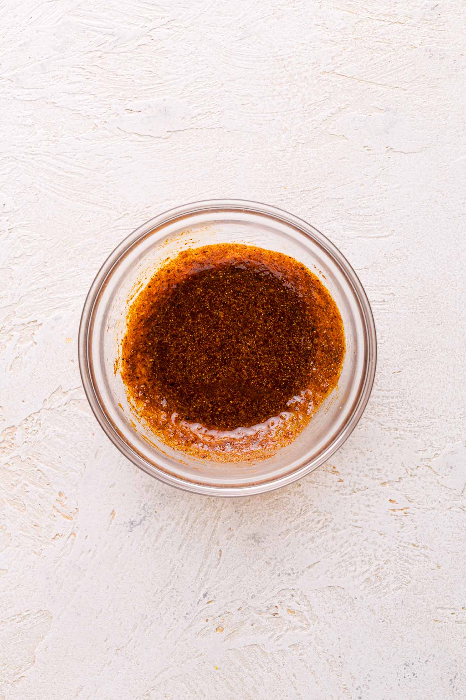 A small glass bowl filled with a dark brown spice mixture on a textured light-colored surface.