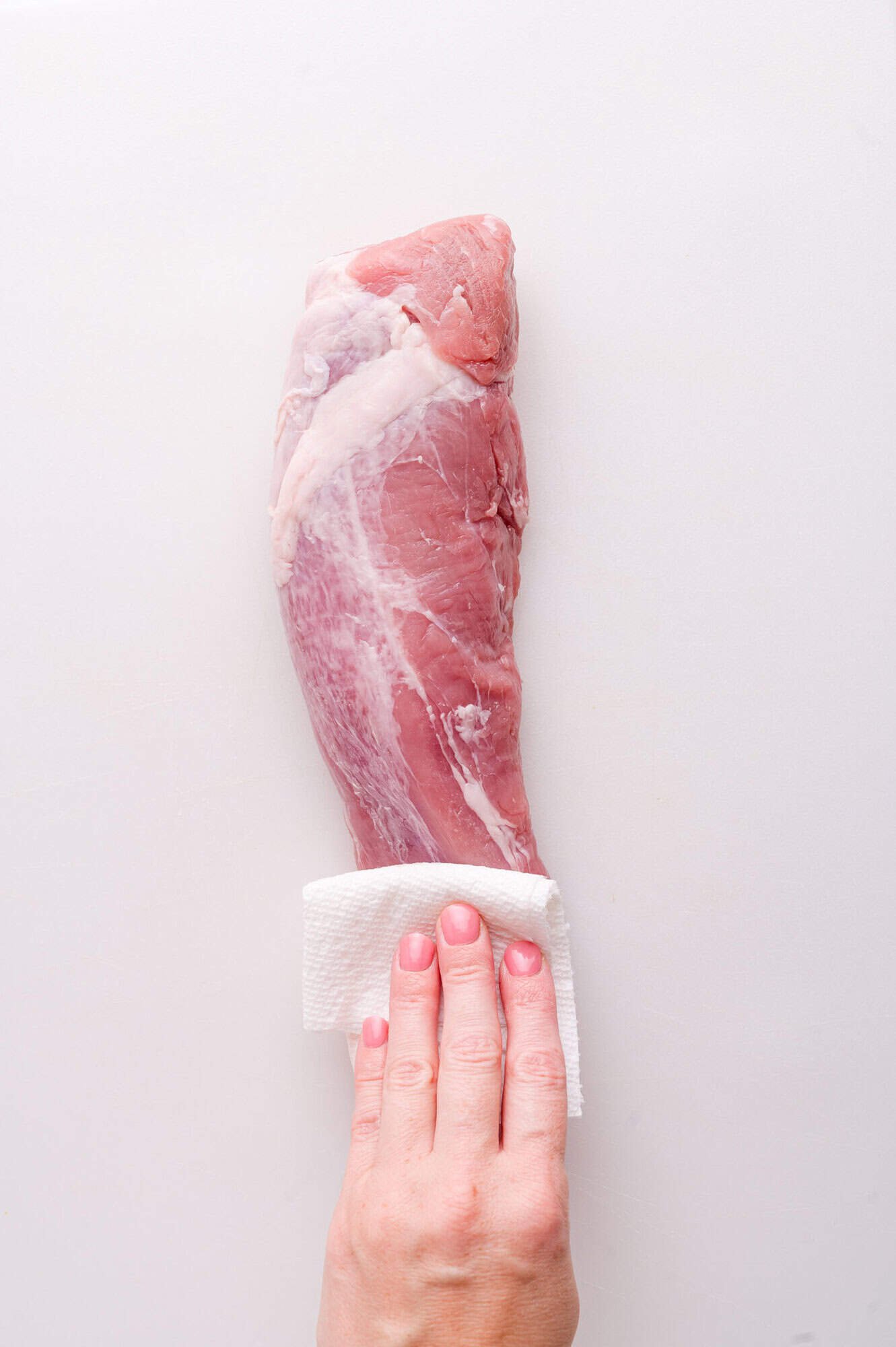 A hand uses a paper towel to pat dry a raw pork tenderloin on a white surface.